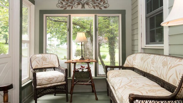 A porch with wicker furniture, a table with a lamp, and a view of greenery outside. - Stay-cation In Charmified Village Guest House! (Aurora)