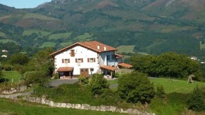 A two-story house with a red-tiled roof, surrounded by lush greenery and hills in the background. - Labakizarra House for 8 people (Baztan)