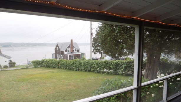 A covered porch with a view of a house across a grassy yard and a body of water.