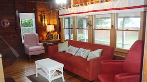 A cozy room with a red sofa, a white coffee table, and a pink armchair. There is a wooden floor and a window with curtains.