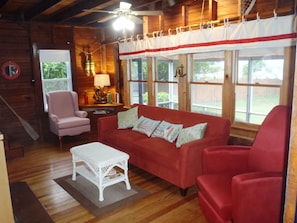 Una habitación acogedora con un sofá rojo, una mesa de centro blanca y un sillón rosa. El suelo es de madera y hay una ventana con cortinas.