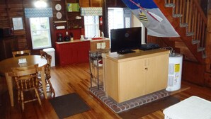 A cozy cabin interior with a wooden dining table, chairs, a kitchen area with a red countertop, a television on a wooden cabinet, and a staircase.