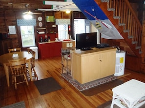 Interior acogedor de una cabaña con una mesa de comedor de madera, sillas, una cocina con encimera roja, un televisor sobre un mueble de madera y una escalera.