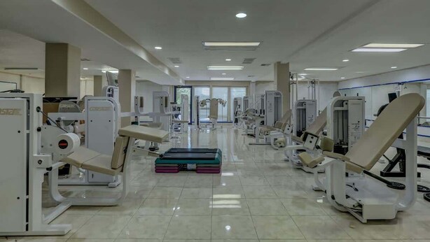A clinical rehabilitation center with various exercise machines, a central bench, and a door leading to another room. - Las Palmas Resort #1511 | All-Inclusive Amenities (Saint George)