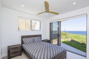 Un dormitorio con cama, ventilador de techo, ventana y puerta corrediza de vidrio que da a un balcón con vistas al mar.