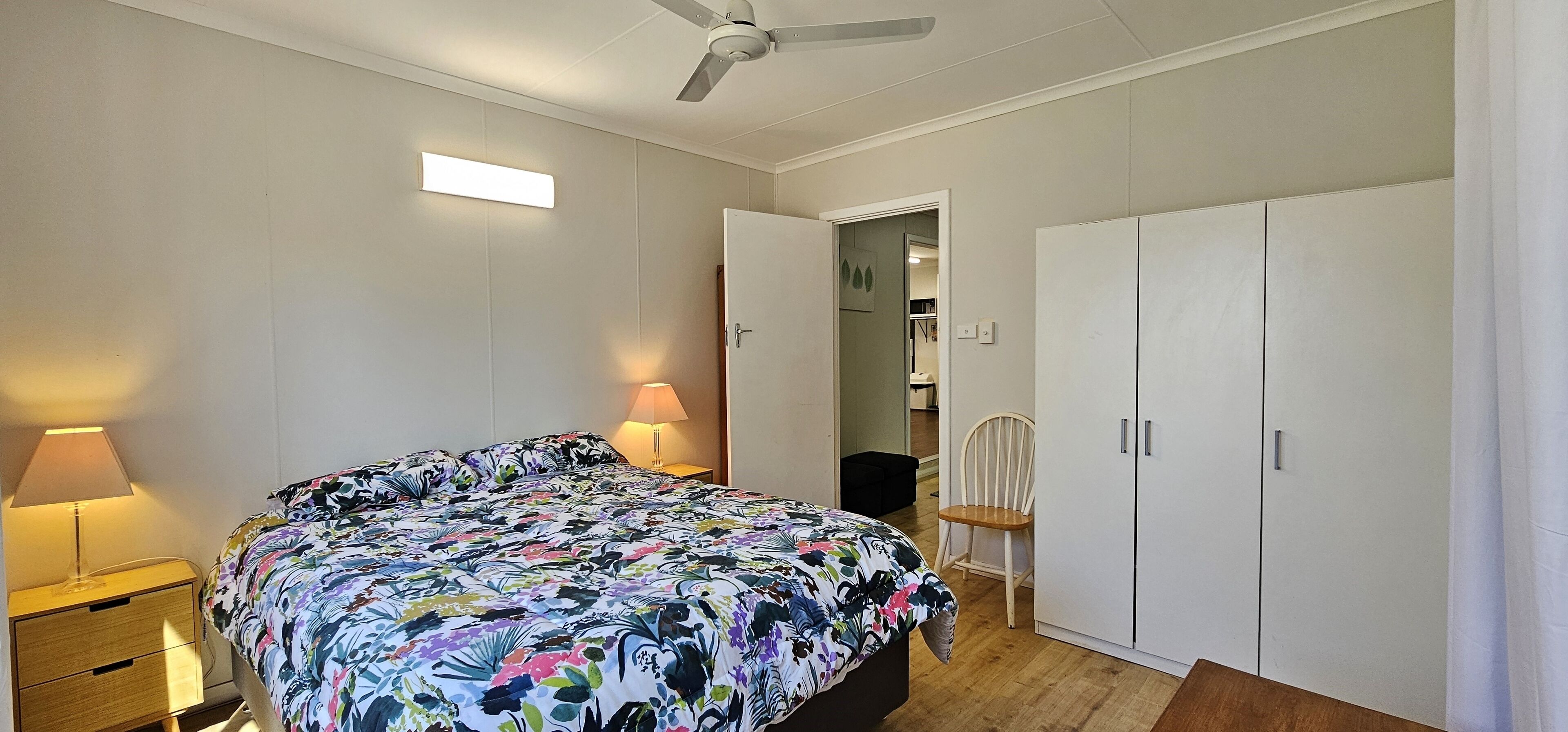 A bedroom with a bed, bedside lamps, a ceiling fan, a wardrobe, and a chair.