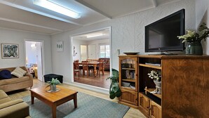 Una sala de estar con sofá, mesa de centro, televisión, estantería y un comedor visible a través de un umbral abierto.