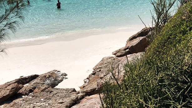Ein Strand mit klarem türkisblauem Wasser, felsigem Ufer und grünen Hügeln im Hintergrund.