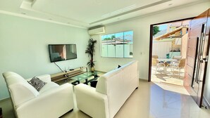 Ein modernes Wohnzimmer mit Glastisch, weißen Sofas, einem Flachbildfernseher und Blick auf die Außenpatio-Terrasse.