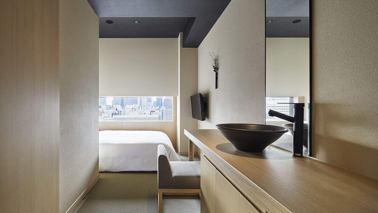 A hotel room with a bed, a desk, a large mirror, a black bowl on the counter, and a view of the city through the window.