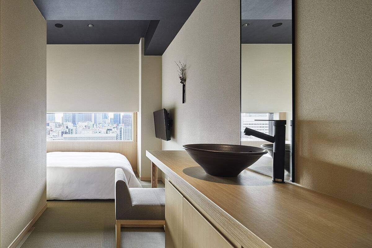 Een hotelkamer met een bed, een bureau, een grote spiegel, een zwarte kom op het aanrecht en uitzicht op de stad.
