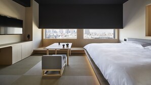 Una habitación de hotel moderna con una cama grande, un televisor de pantalla plana, una mesa de comedor y vistas a la ciudad a través de amplias ventanas.