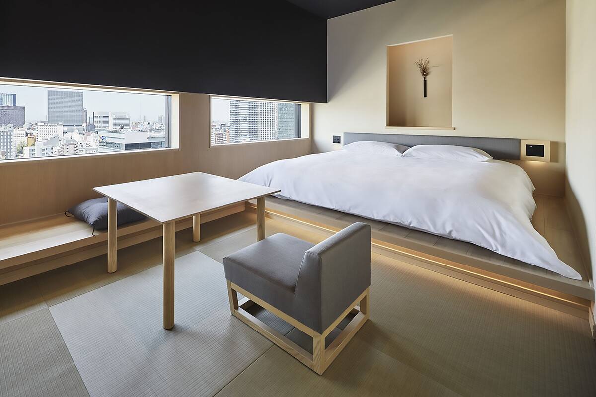 Een moderne hotelkamer met een bed, een klein tafeltje, een bankje en uitzicht op de stad.