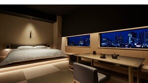 Une chambre d’hôtel moderne, équipée d’un lit, d’un bureau et d’une chaise. Elle possède deux fenêtres donnant sur la ville.