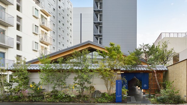 Un bâtiment traditionnel japonais, doté d’un toit en bois et d’une porte d’entrée bleue, entouré de verdure et situé à côté d’immeubles modernes de grande hauteur.