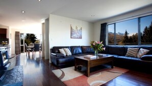 A modern living room with a dark leather sofa, a wooden coffee table, and a large window offering a view of a mountainous landscape. - Spectacular Views , Large & Modern , Perfect For Families & Large Groups (Queenstown)