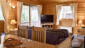 Interior acogedor de una cabaña con techo de madera, un televisor sobre un mueble de madera, un sofá de cuero, una mesa de comedor con sillas y una cesta.