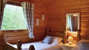 Una cabaña con dos camas de madera, una ventana con cortinas, una lámpara de noche y cuadros enmarcados en la pared.
