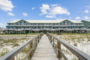 Et hotel ved stranden med en træpromenade, der fører til indgangen, flere balkoner og en klar himmel.