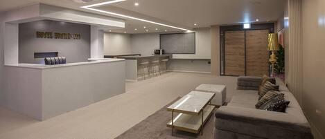 A modern hotel lobby with a reception desk, seating area, and a kitchen in the background.
