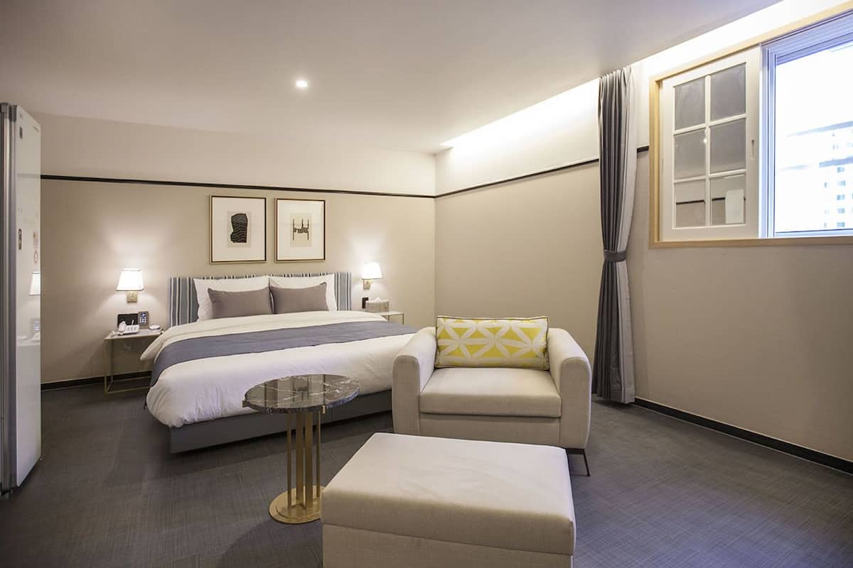 A hotel room with a bed, a chair, a small table, and a window with curtains.