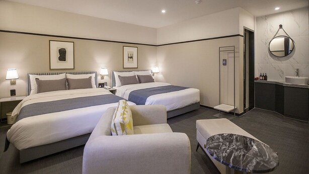 Habitación de hotel con dos camas, una zona de estar, una mesita, un espejo y un baño con lavamanos e inodoro.