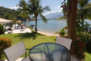 Un patio con mesa y sillas de vidrio, con vista a una playa y un lago con barcos.
