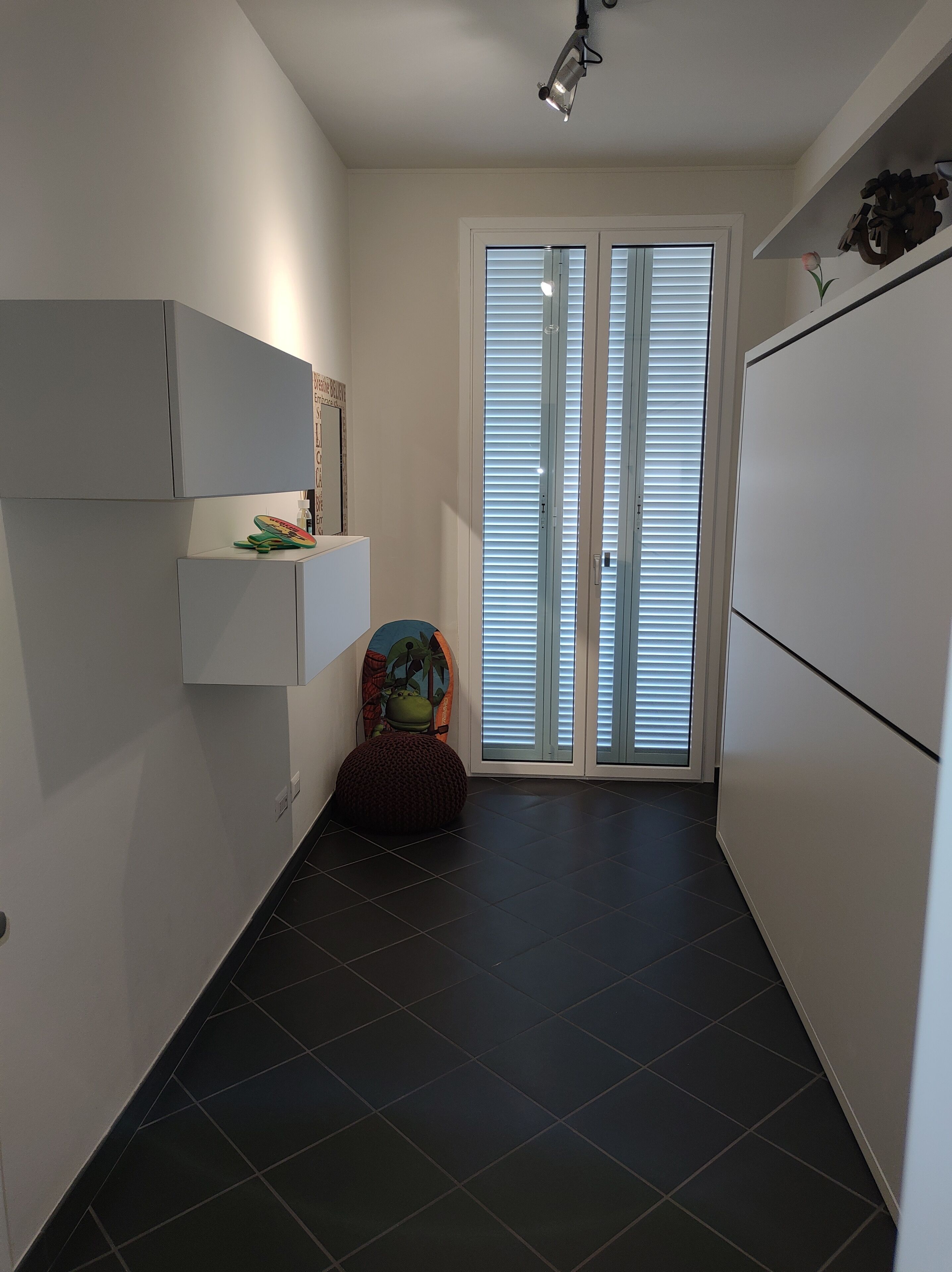 En smal hall med ett mörkt klinkergolv, vita väggar och en dörr med persienner.