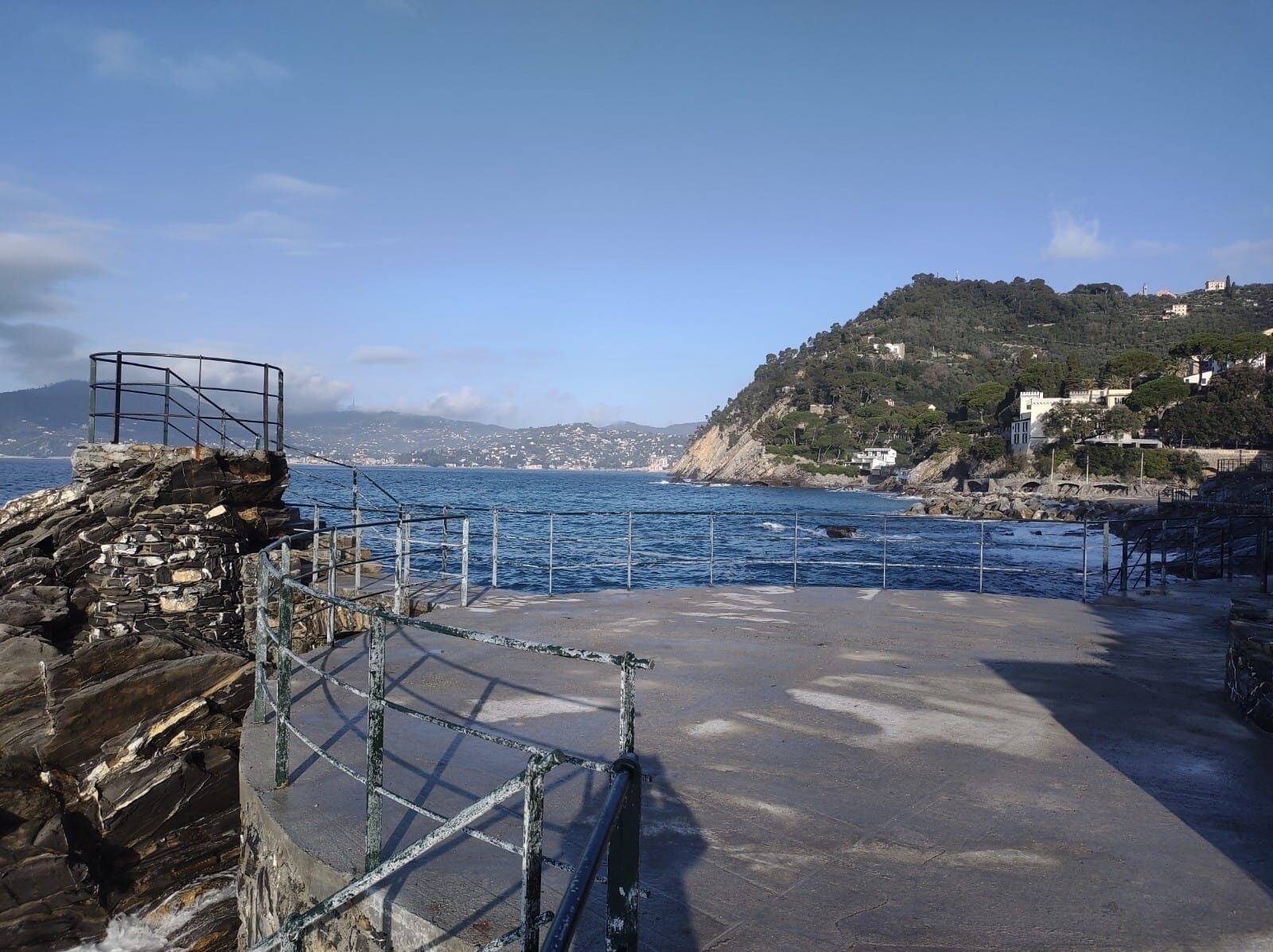 A coastal area with a rocky pier, a metal railing, and a view of the sea and distant hills.