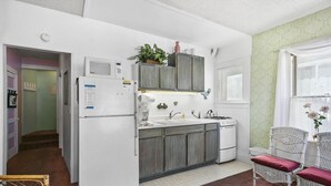 A compact kitchen with a refrigerator, sink, and wooden cabinets. There is a wicker chair with a cushion and a small table with a vase of flowers. - 216WA: Budget-friendly Apartment in Victorian Triplex, 2 Blocks to the Beach + W (Avalon)