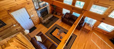 A cabin interior with a stone fireplace, wooden floors, a brown leather sofa, and multiple windows.