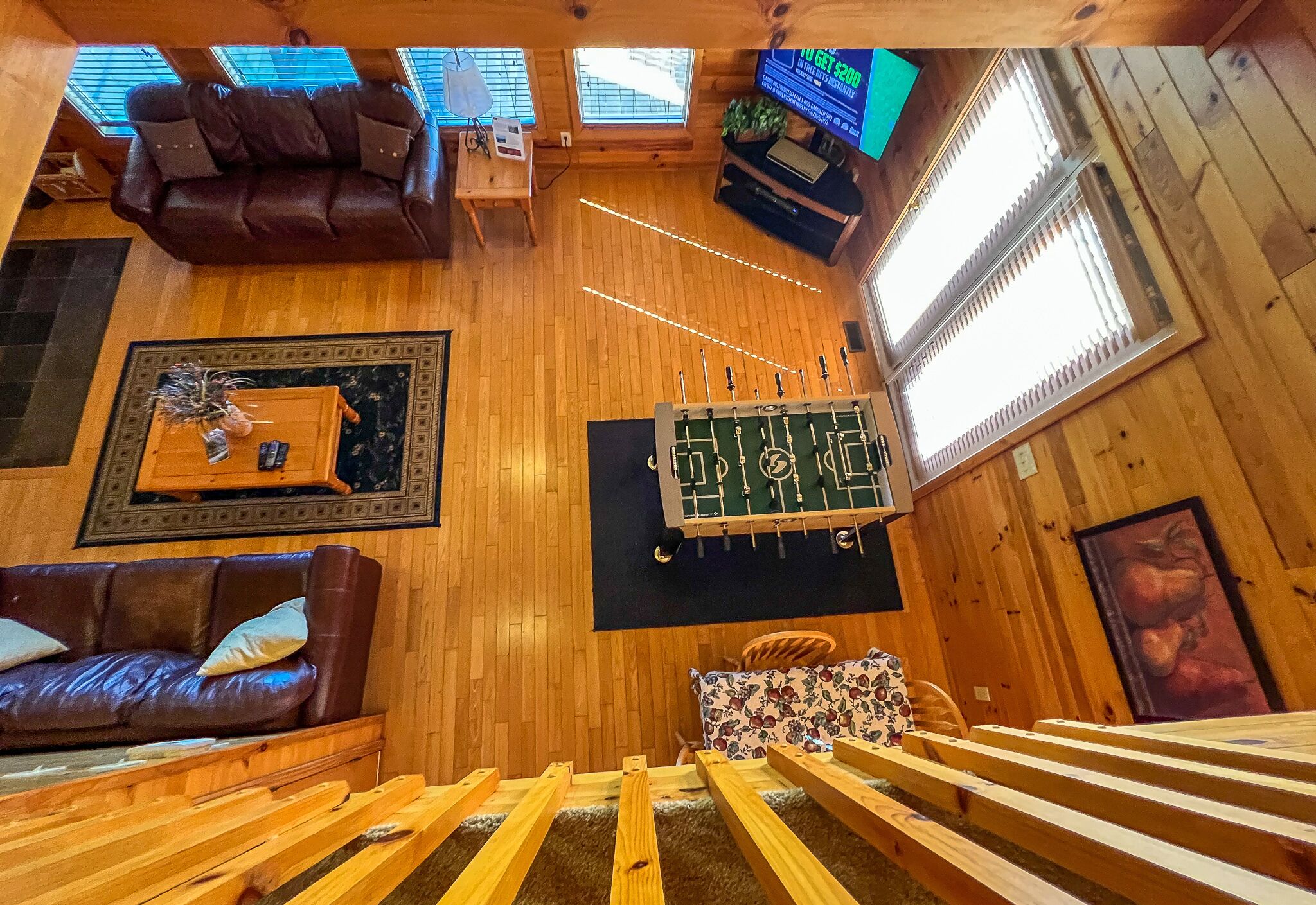 A cozy room with a wooden floor, a sofa, a coffee table, a foosball table, a TV, and a window with blinds.