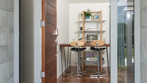 A room with a wooden door, a small table with two chairs, a shelf with books and a plant, and a view through a glass door. - Spacious Modern Two-Bedroom Apartment (Tamahere)