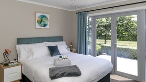 A modern bedroom with a large bed, a nightstand, a bedside table, and a view of a garden through a sliding glass door. - Spacious Modern Two-Bedroom Apartment (Tamahere)