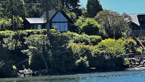 A lakeside property with a house featuring a large bay window and a metal roof, surrounded by dense greenery and a rocky shoreline. - Private Oceanfront Cabin with Hot Tub / Doc Holiday Cabin (Ucluelet)