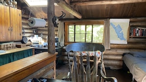 A rustic cabin interior with wooden walls, a dining table, a bench, a bed, and a map on the wall.