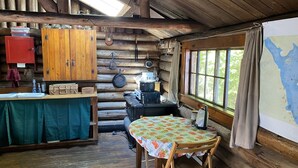 A rustic cabin interior with a wooden table and chairs, a kitchen area with a stove, and a window with curtains.