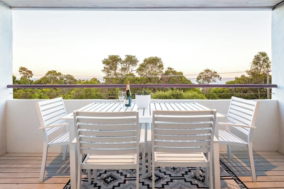 Un balcon avec une table et des chaises, une bouteille de champagne et une vue sur les arbres et un ciel dégagé.