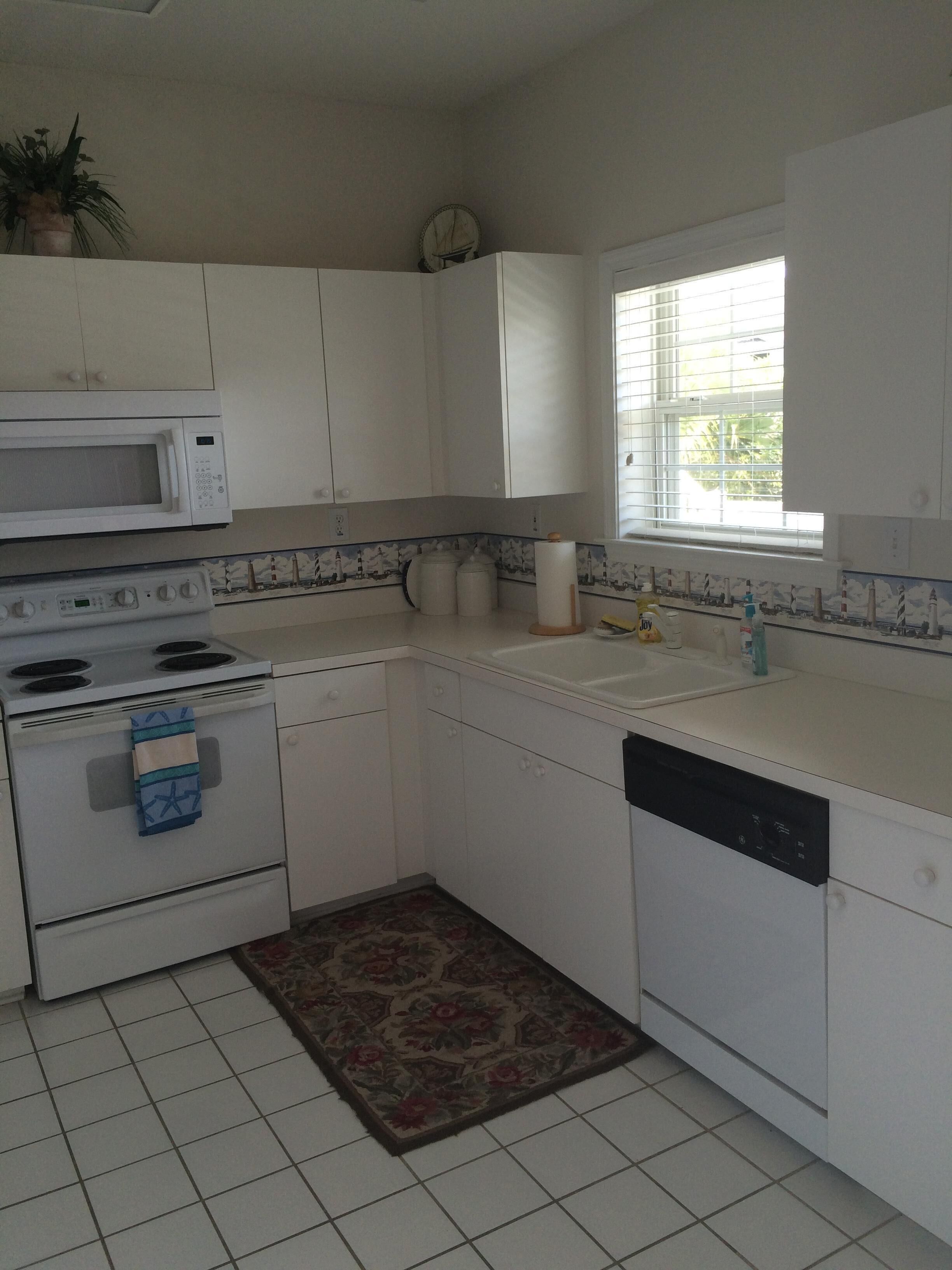 A compact kitchen with white cabinets, a microwave, stove, and dishwasher. There's a window with blinds and a decorative rug on the floor.