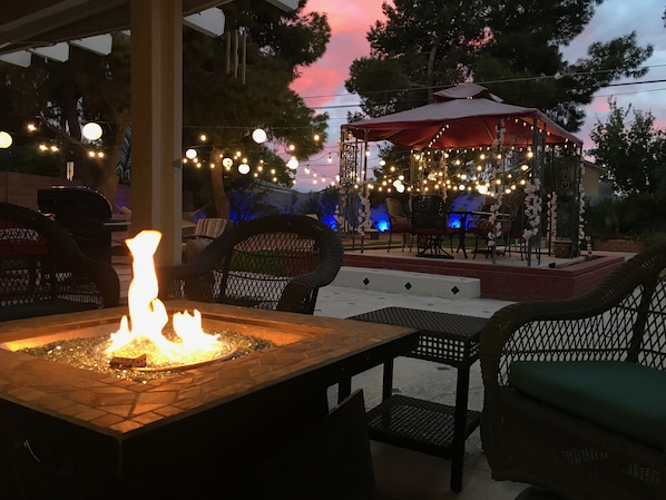Un patio al aire libre con zona de fuego, asientos y luces colgantes.