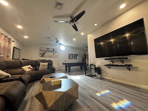 Una sala de estar con un sofá de cuero marrón, una mesa de centro de madera, un televisor de pantalla plana montado en la pared y un ventilador de techo.