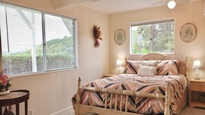Une chambre à coucher comprenant un lit, deux tables de chevet, une fenêtre avec des stores, un élément de décoration fixé au mur, et une petite table sur laquelle se trouvent un vase et une nichoir.