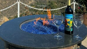 Une table avec un foyer, agrémentée d’une bouteille et de verres, entourée d’une clôture en treillis métallique, et offrant une vue sur un paysage montagneux.