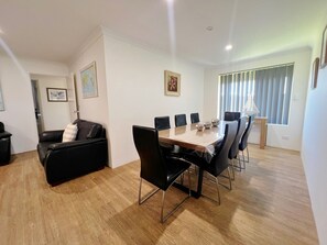 Un salon moderne avec un canapé en cuir noir, une table à manger en bois et six chaises, ainsi qu’une grande fenêtre équipée de stores.