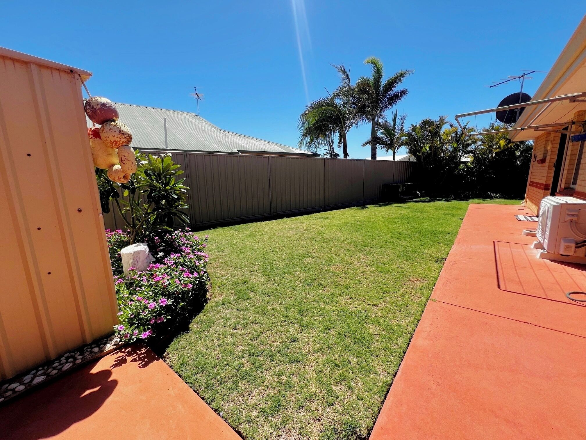 A backyard with a lawn, a concrete pathway, a small garden with pink flowers, and a metal fence.