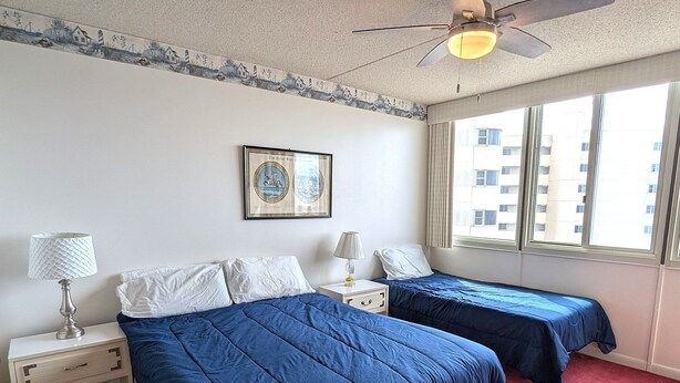 A hotel room with two beds, a ceiling fan, a window with a view of buildings, and a framed picture on the wall.