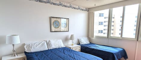 Hotelkamer met twee bedden, een plafondventilator, een raam met uitzicht op gebouwen en een ingelijst schilderij aan de muur.