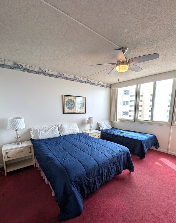 Ein Hotelzimmer mit zwei Betten, einem Deckenventilator, einem Fenster mit Blick auf Gebäude und einem gerahmten Bild an der Wand.