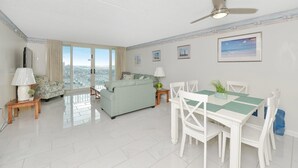 A spacious living area with a sofa, dining table, and chairs, a ceiling fan, and a city view through large windows.