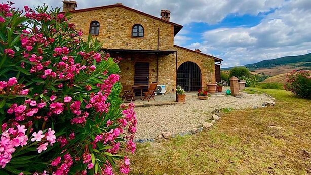 A stone house with a large arched entrance, surrounded by a gravel driveway and flowering plants.